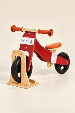 A red and wooden 2-in-1 Mini Balance Bike features a sturdy design with an orange seat and a wooden stand for storage.