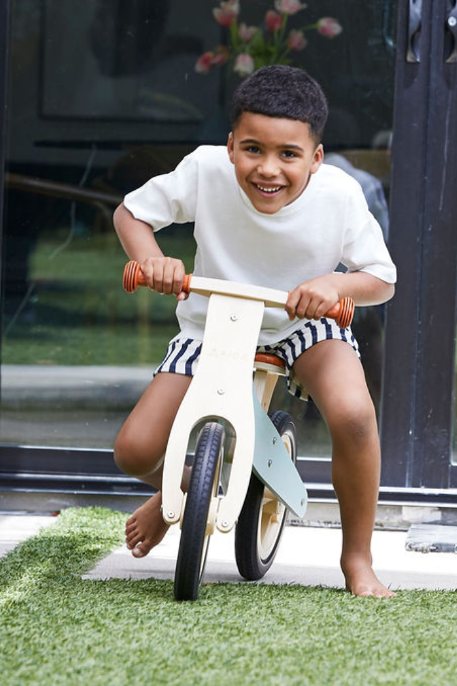 A young boy rides a wooden balance bike on a grassy area, smiling joyfully. The bike features a light wooden frame with red handlebars, perfect for children aged 3 to.