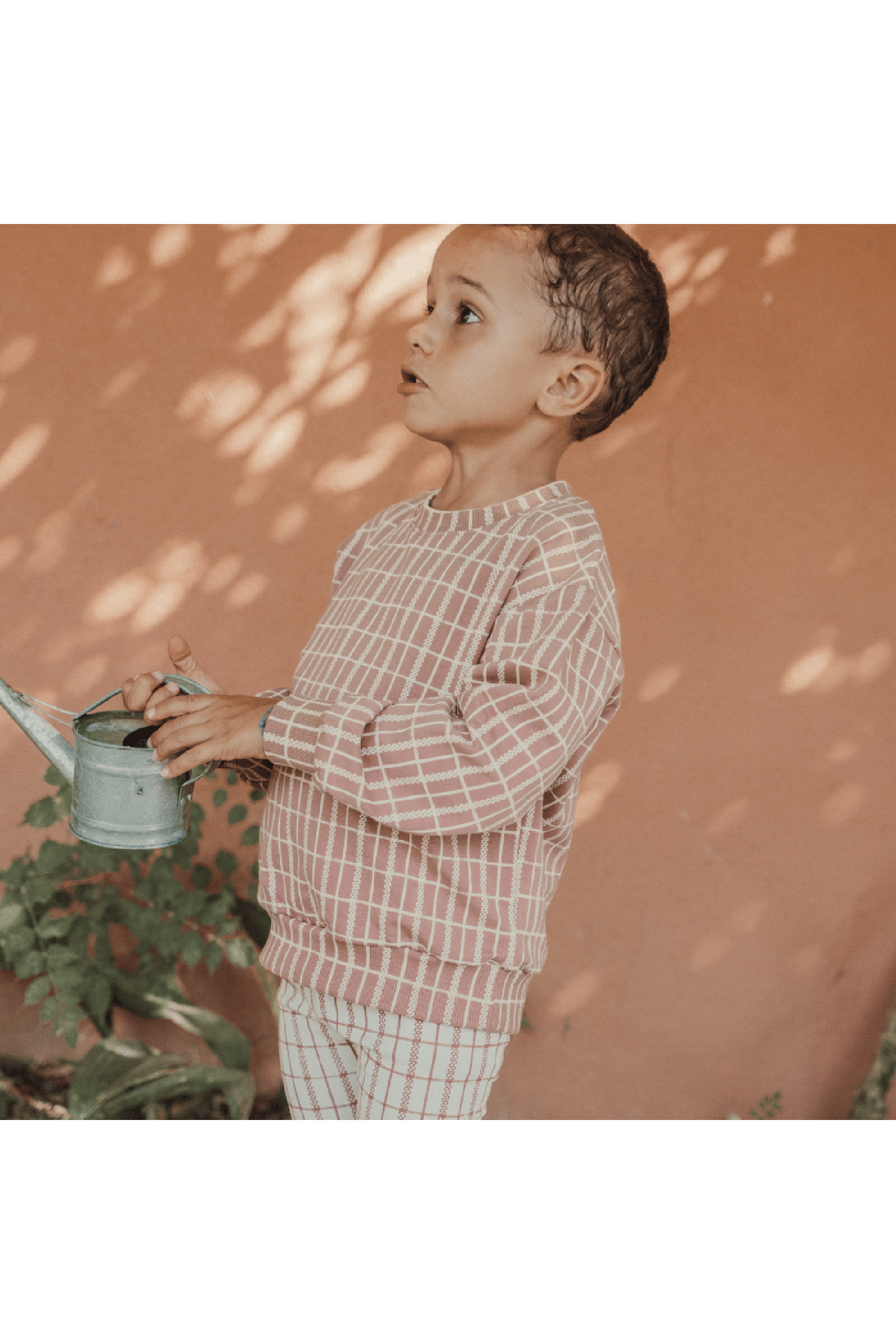 A young child stands against a warm orange wall, wearing a mauve check printed sweatshirt. The cozy cotton blend garment features a relaxed fit, perfect for outdoor play or lounging.