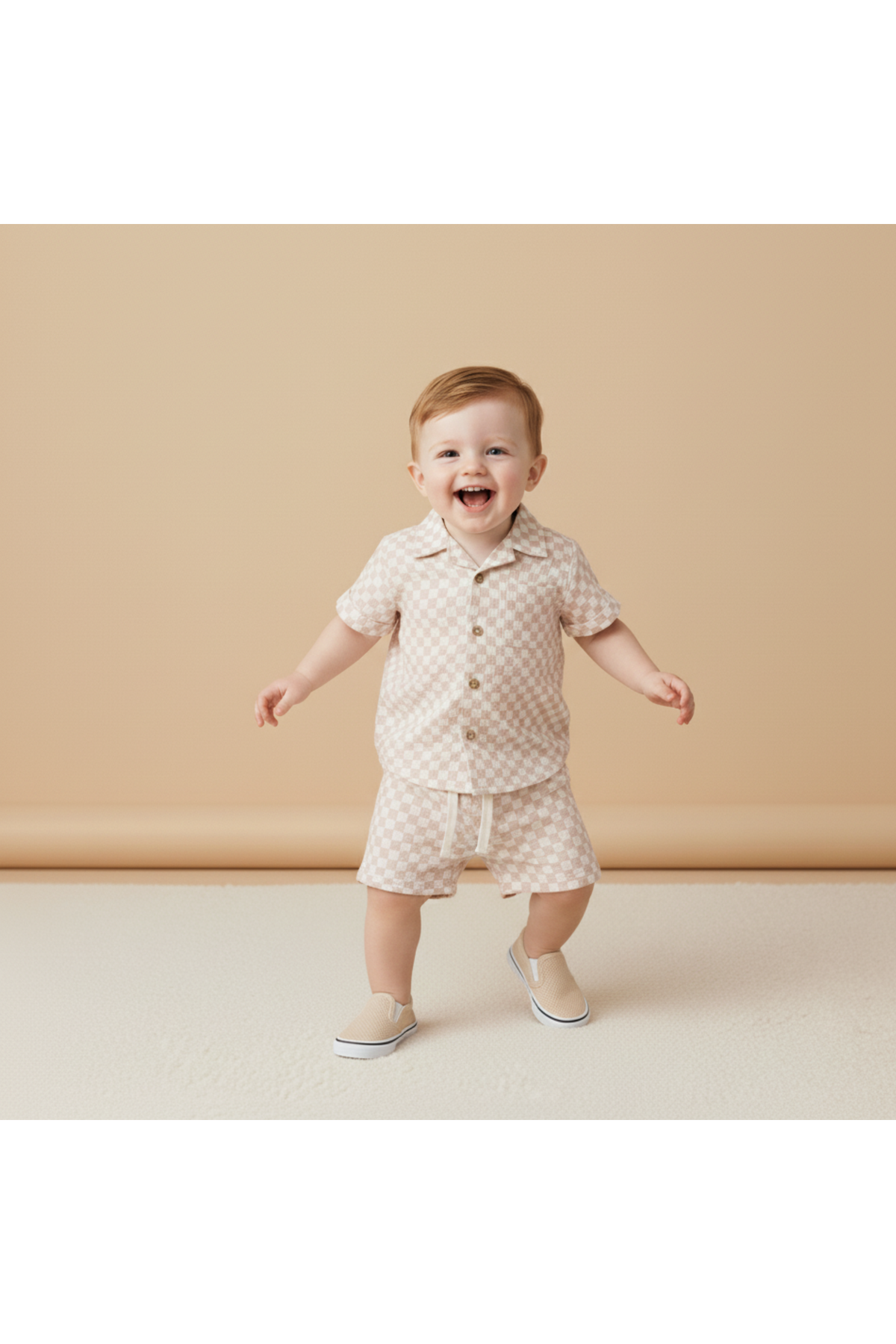 A cheerful toddler wearing a light brick checkered organic cotton shirt and shorts set smiles while standing on a textured surface against a soft, neutral background. Perfect for summer adventures!