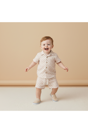 A cheerful toddler wearing a light brick checkered organic cotton shirt and shorts set smiles while standing on a textured surface against a soft, neutral background. Perfect for summer adventures!