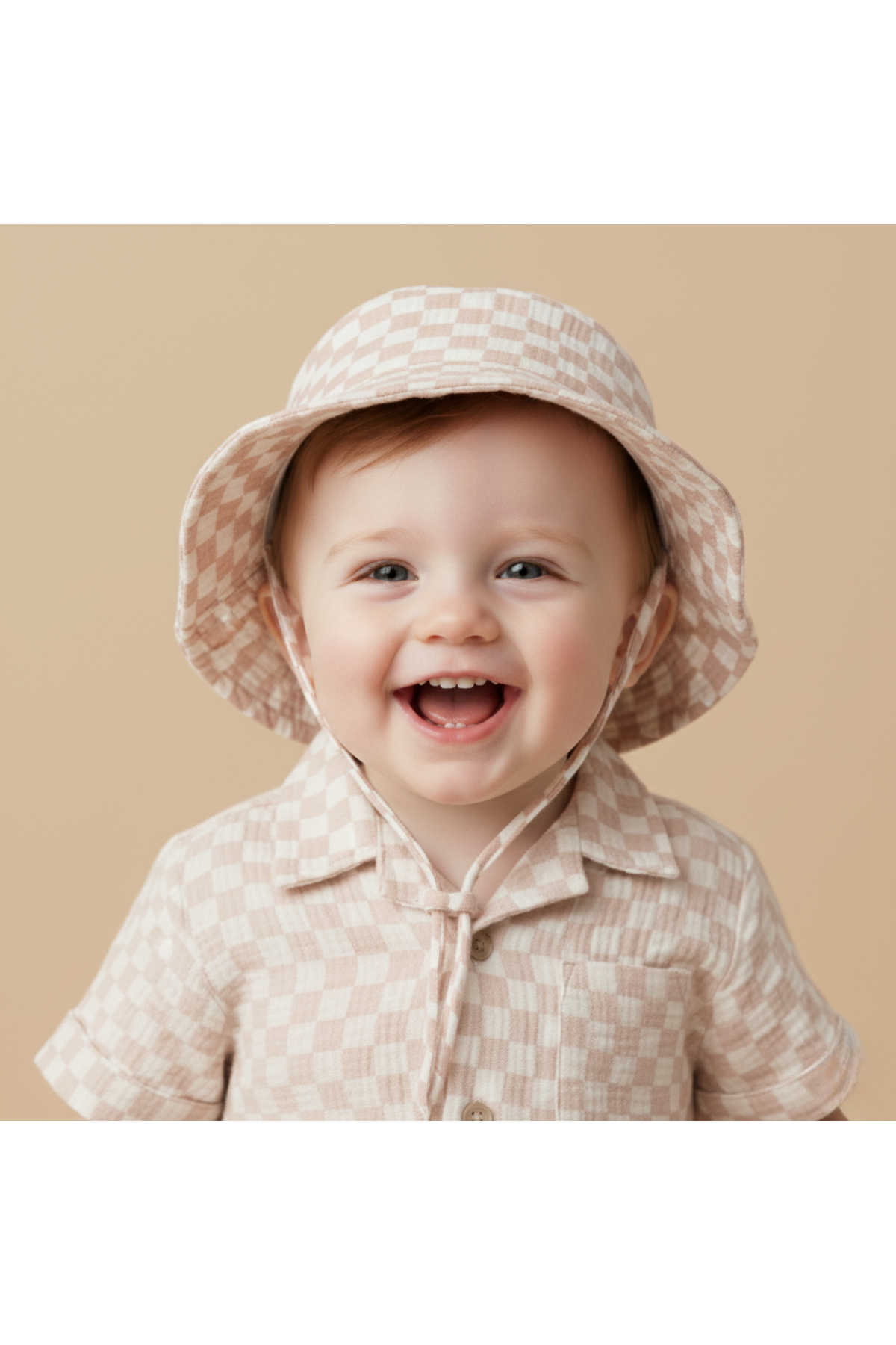 A smiling toddler wears a light brick check-patterned bucket hat paired with a matching short-sleeve button-up shirt. The soft, organic cotton hat offers sun protection and comfort for outdoor play.