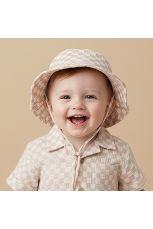 A smiling toddler wears a light brick check-patterned bucket hat paired with a matching short-sleeve button-up shirt. The soft, organic cotton hat offers sun protection and comfort for outdoor play.