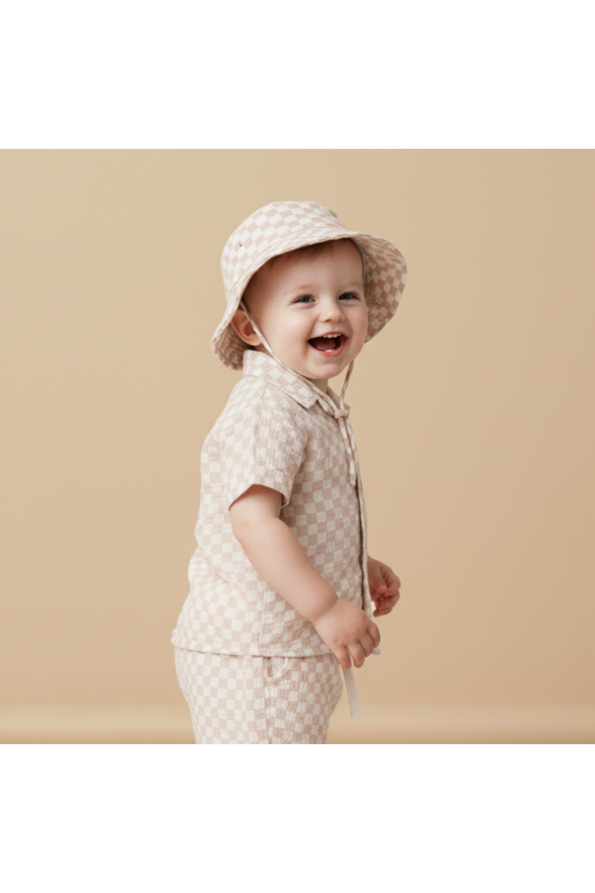 A smiling toddler wearing a light-colored checkered bucket hat and matching short-sleeve shirt stands against a soft beige background, showcasing a playful, comfortable style perfect for sunny outings.