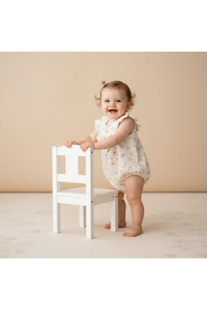 A joyful baby stands beside a small white chair, wearing a sleeveless, blush floral romper. The soft fabric features ruffles, emphasizing its playful and comfortable design for little ones.