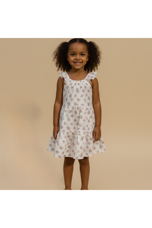 A young girl wearing a blush floral ruffle tiered dress stands smiling against a neutral background. The dress features delicate ruffles and a lightweight, flowy design, perfect for warm weather.