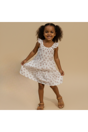 A young girl smiles while twirling in a blush floral tiered dress with ruffles. The lightweight, organic cotton fabric enhances her playful, carefree look against a neutral background.