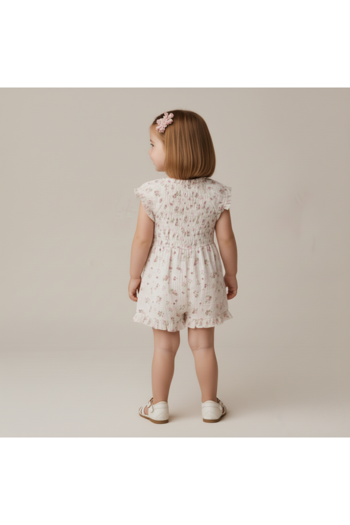 A young child wears a blush floral organic smocked jumpsuit, featuring delicate smocking and short sleeves. The soft muslin cotton fabric is perfect for warm, casual outings.