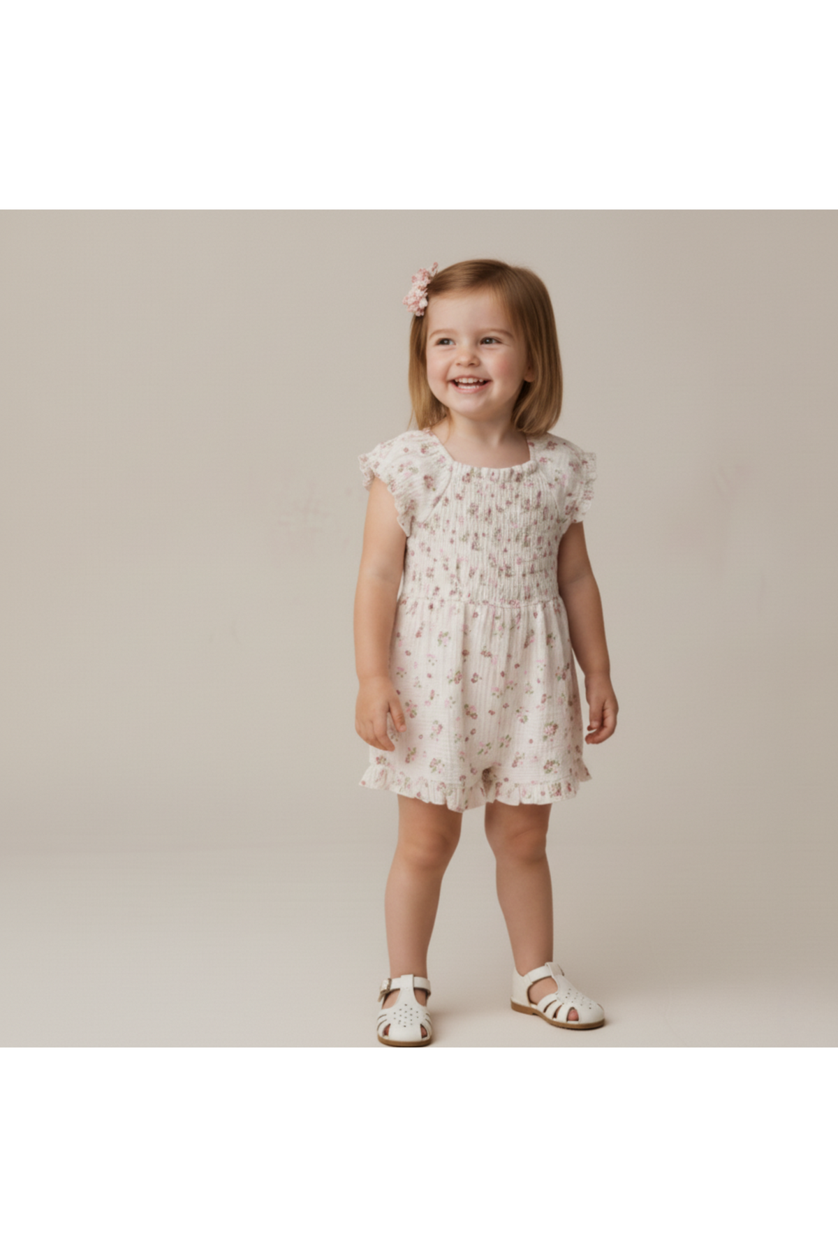 A smiling young girl wears a soft, blush floral jumpsuit with smocked detailing. The outfit features short sleeves and a comfortable fit, perfect for warm, playful days.