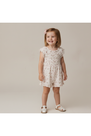 A smiling young girl wears a soft, blush floral jumpsuit with smocked detailing. The outfit features short sleeves and a comfortable fit, perfect for warm, playful days.