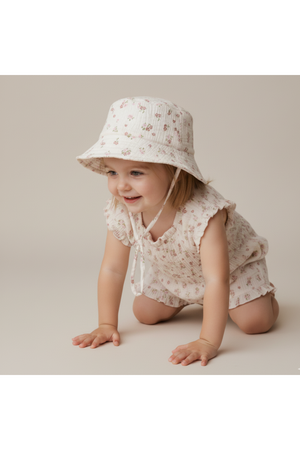 A smiling toddler crawls on a neutral background, wearing a blush floral organic bucket sun hat and a matching floral outfit.