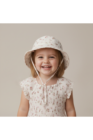 A cheerful young girl wears a blush floral bucket sun hat made from soft organic cotton. The hat features a wide brim and is styled with a lightweight, comfortable design.