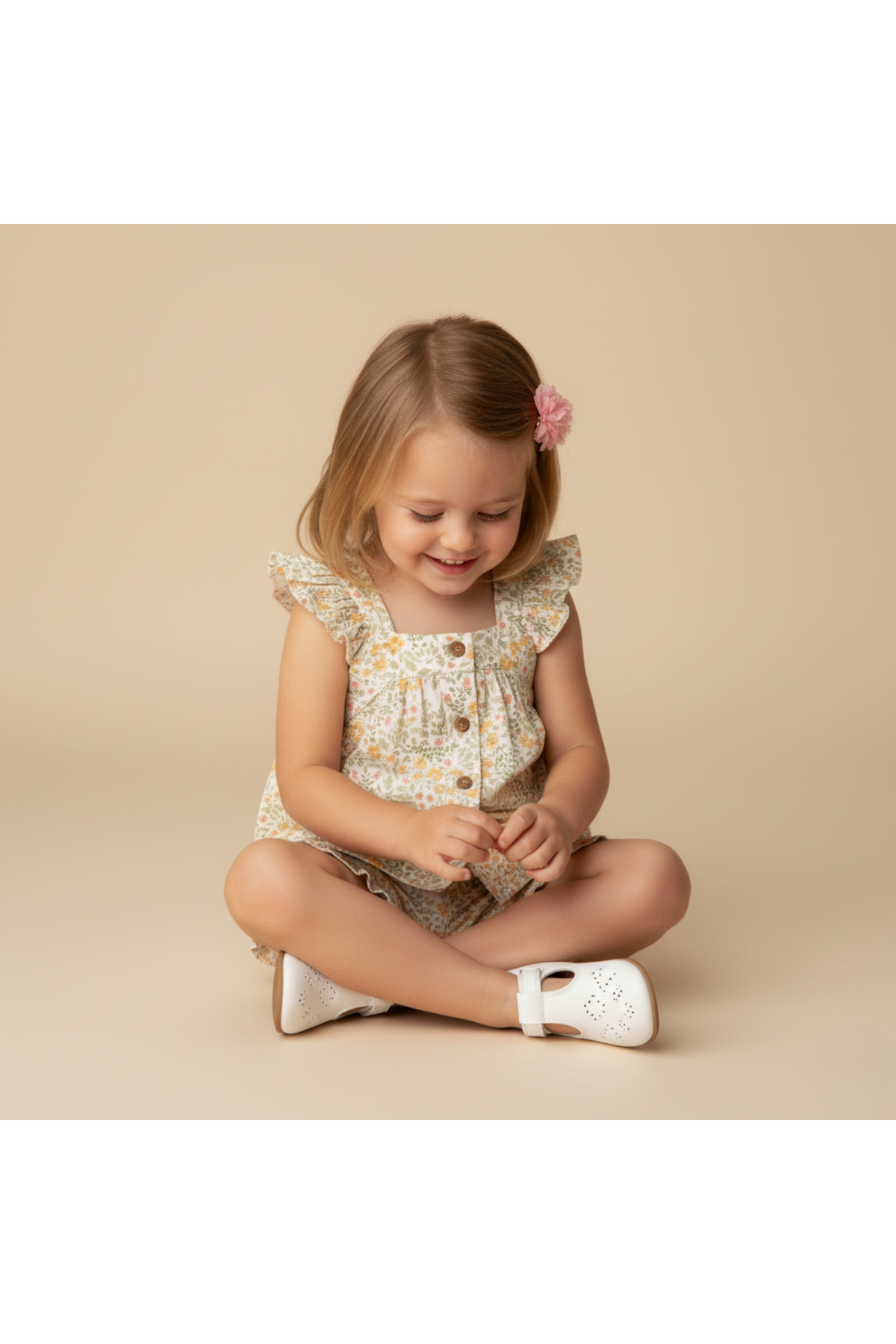 A young girl sits on the floor, wearing a floral flutter top and shorts set made from organic cotton. She smiles while playing, showcasing a cheerful summer vibe.