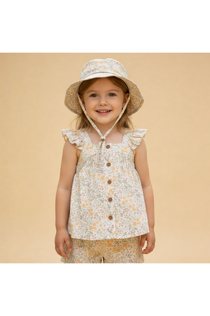 A smiling child wears a floral-patterned top with ruffled sleeves and a matching bucket sun hat. The outfit features soft, organic cotton, perfect for sunny days and outdoor play.