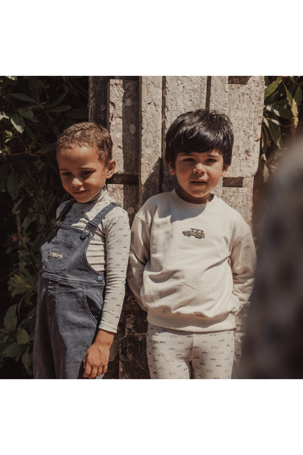 Two children stand against a stone wall, one wearing a denim overall and the other in a light sweatshirt. The setting features greenery, highlighting a playful, outdoor vibe.