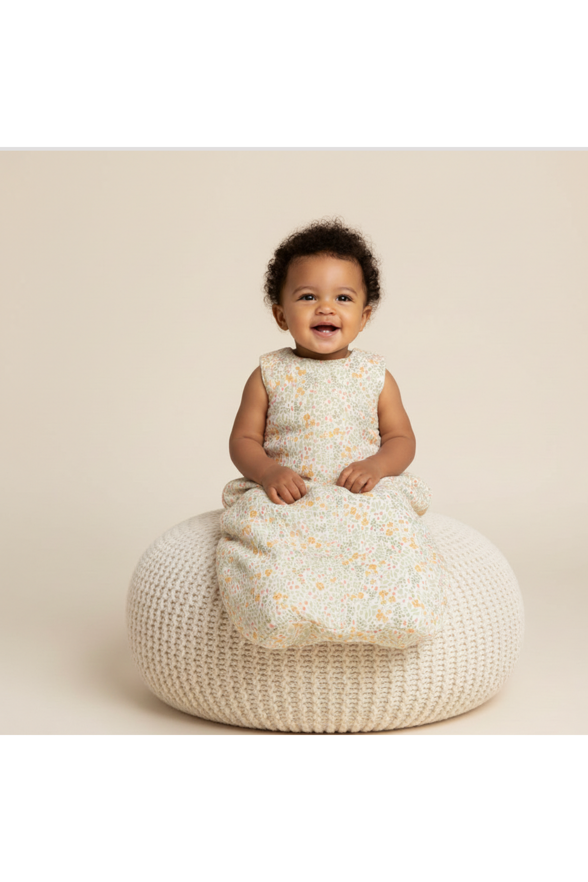 A smiling toddler sits on a textured, cream-colored pouf, wearing a floral-patterned organic cotton wearable blanket. The soft, breathable fabric offers comfort and style for peaceful slumbers.