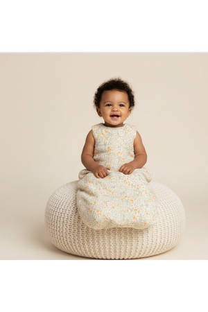 A smiling toddler sits on a textured, cream-colored pouf, wearing a floral-patterned organic cotton wearable blanket. The soft, breathable fabric offers comfort and style for peaceful slumbers.