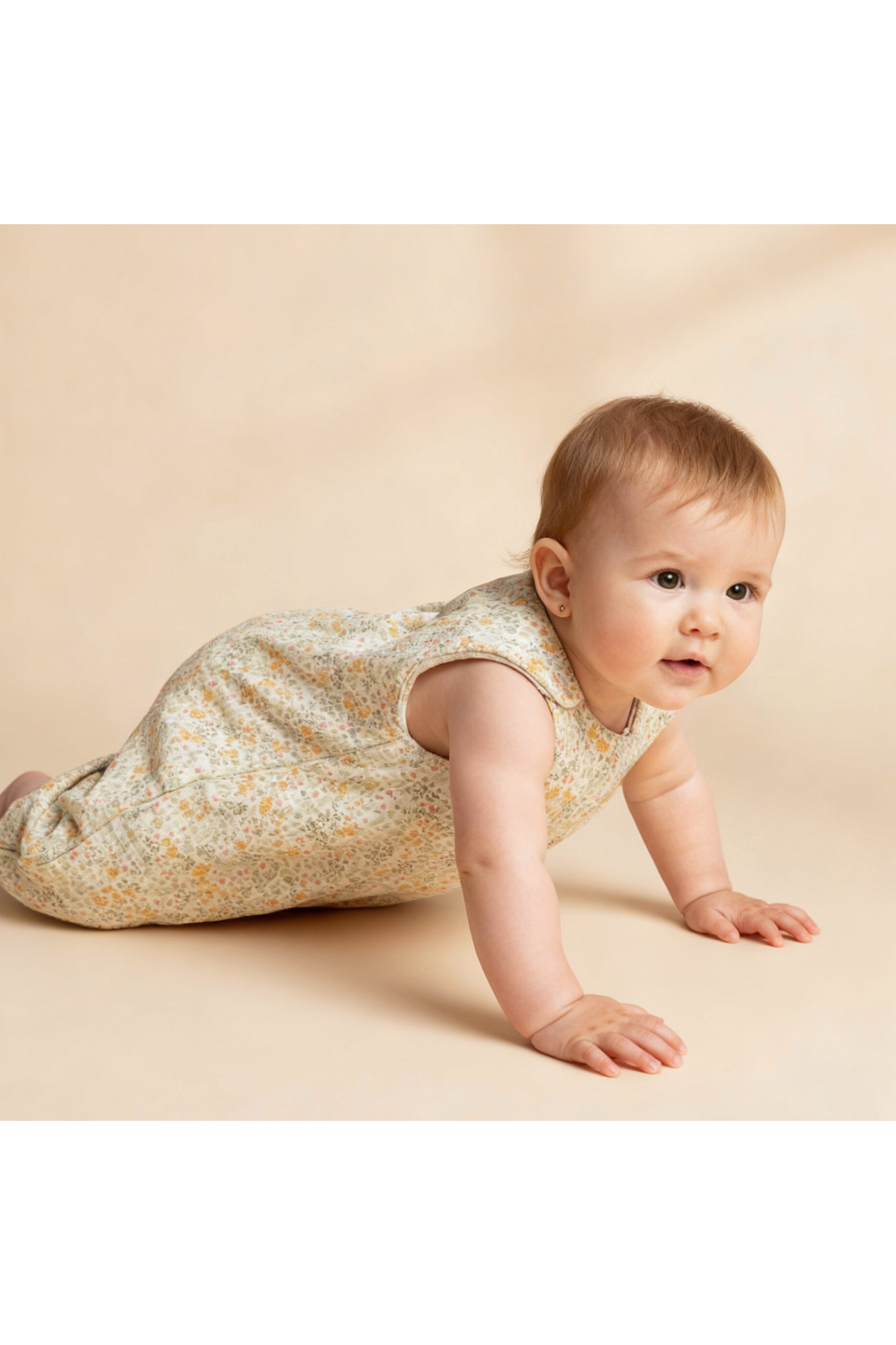 A baby crawls on a soft, neutral backdrop, wearing a floral-patterned organic cotton wearable blanket. The blanket features a sleeveless design and is designed for comfort and coziness.
