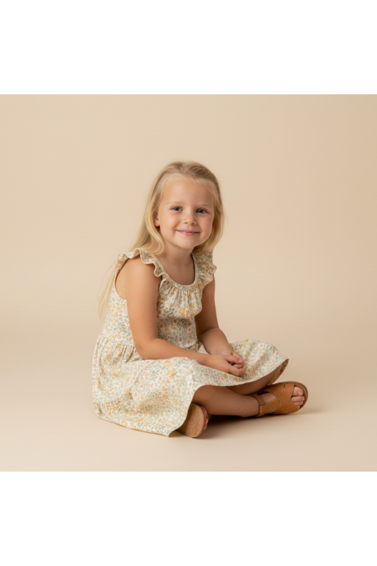 A young girl sits on a light beige background, wearing an organic cotton tiered dress with a floral pattern and ruffled sleeves, complemented by brown sandals.