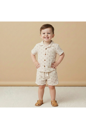 A smiling child stands confidently in a cream-colored organic cotton shirt and shorts set adorned with small brown stars. The background is a soft beige, enhancing the outfits playful nature.