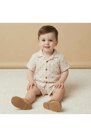 A smiling toddler sits on a soft, light-colored surface, wearing a beige short-sleeve shirt and matching shorts with a subtle print.