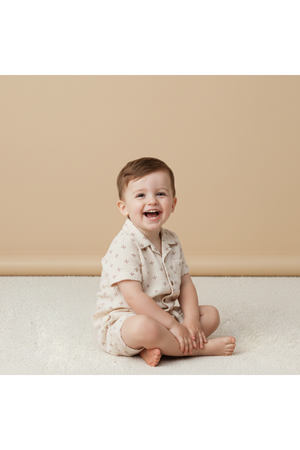 A smiling toddler sits on a soft, light-colored surface, wearing a beige organic cotton shirt and shorts set featuring a subtle cross pattern. The background is a warm, neutral tone.