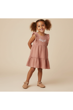 A young girl stands smiling in a tiered, clay-colored dress with delicate embroidery, featuring ruffled sleeves. The soft muslin cotton fabric offers a playful and comfortable fit for sunny days.
