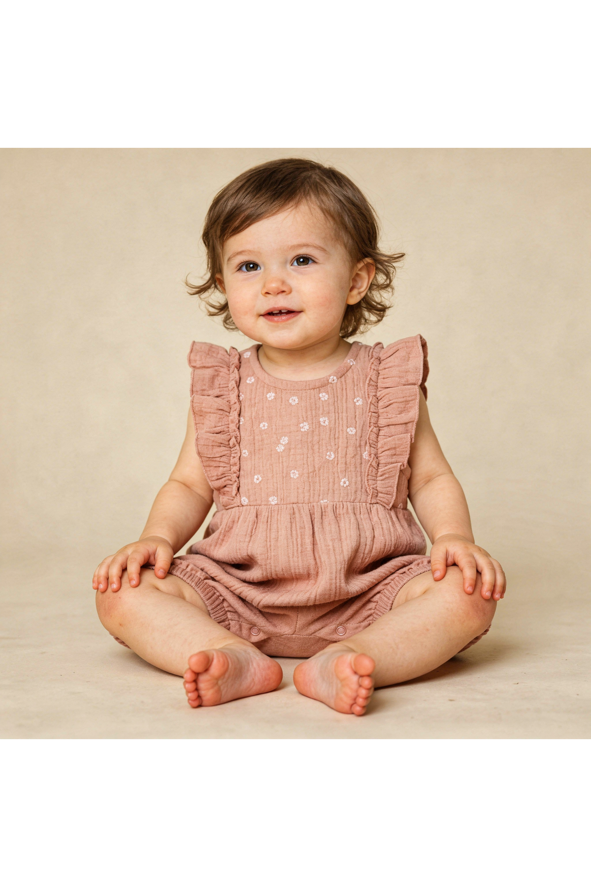 A toddler sits on a neutral background, wearing a clay-colored organic cotton romper with ruffle accents and delicate embroidery. The child has curly hair and a cheerful expression.