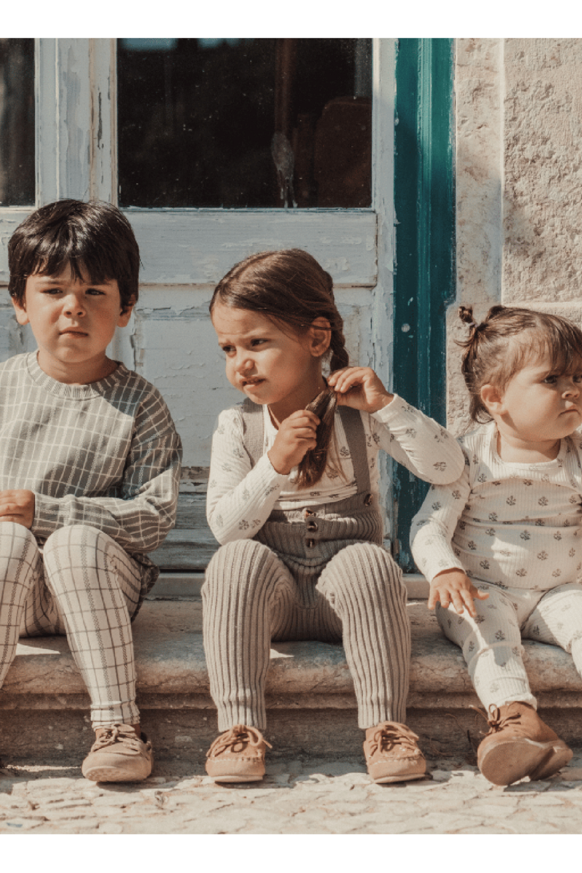 Three children sit on stone steps in front of a colorful door. They wear cozy, patterned outfits in soft, neutral colors, showcasing a playful and comfortable style.