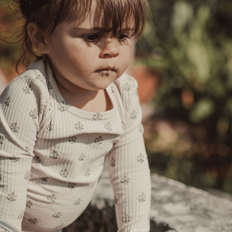 A young child sits outdoors, wearing a cream-colored long-sleeve shirt with a subtle floral print. The soft cotton fabric is gentle against the skin, perfect for comfortable play.