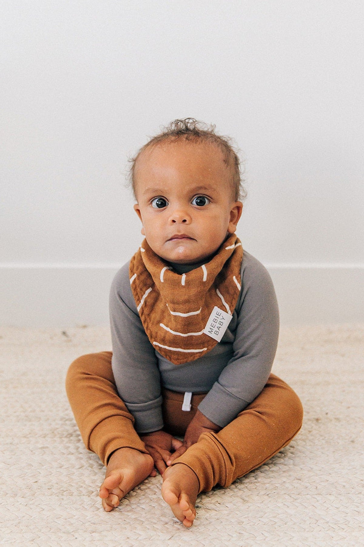 A baby wearing a mustard-colored Mebie Baby muslin bib and matching pants, sitting on a woven rug.