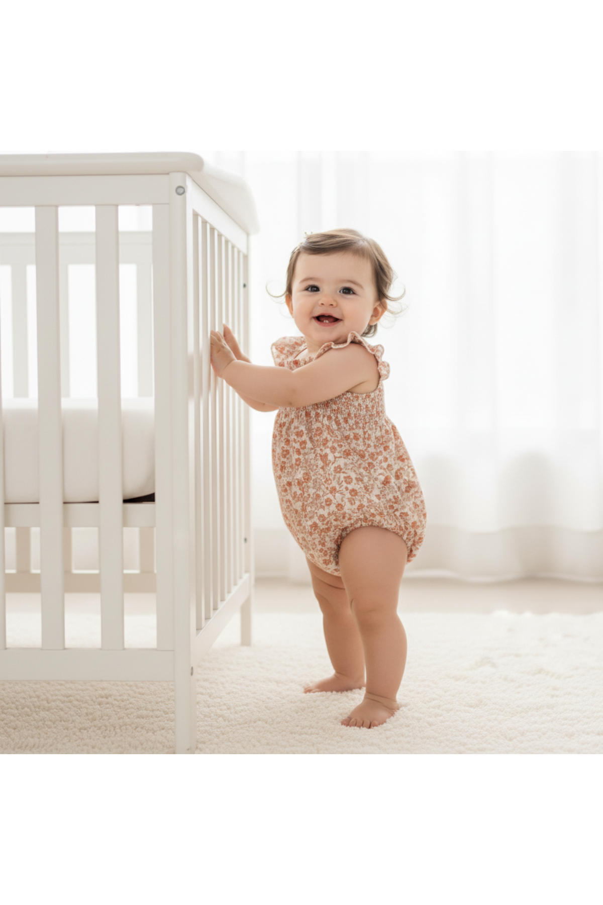 A smiling baby stands next to a white crib, wearing a soft, floral-patterned organic bubble romper in warm tones. The bright, airy room features natural light filtering through sheer curtains.