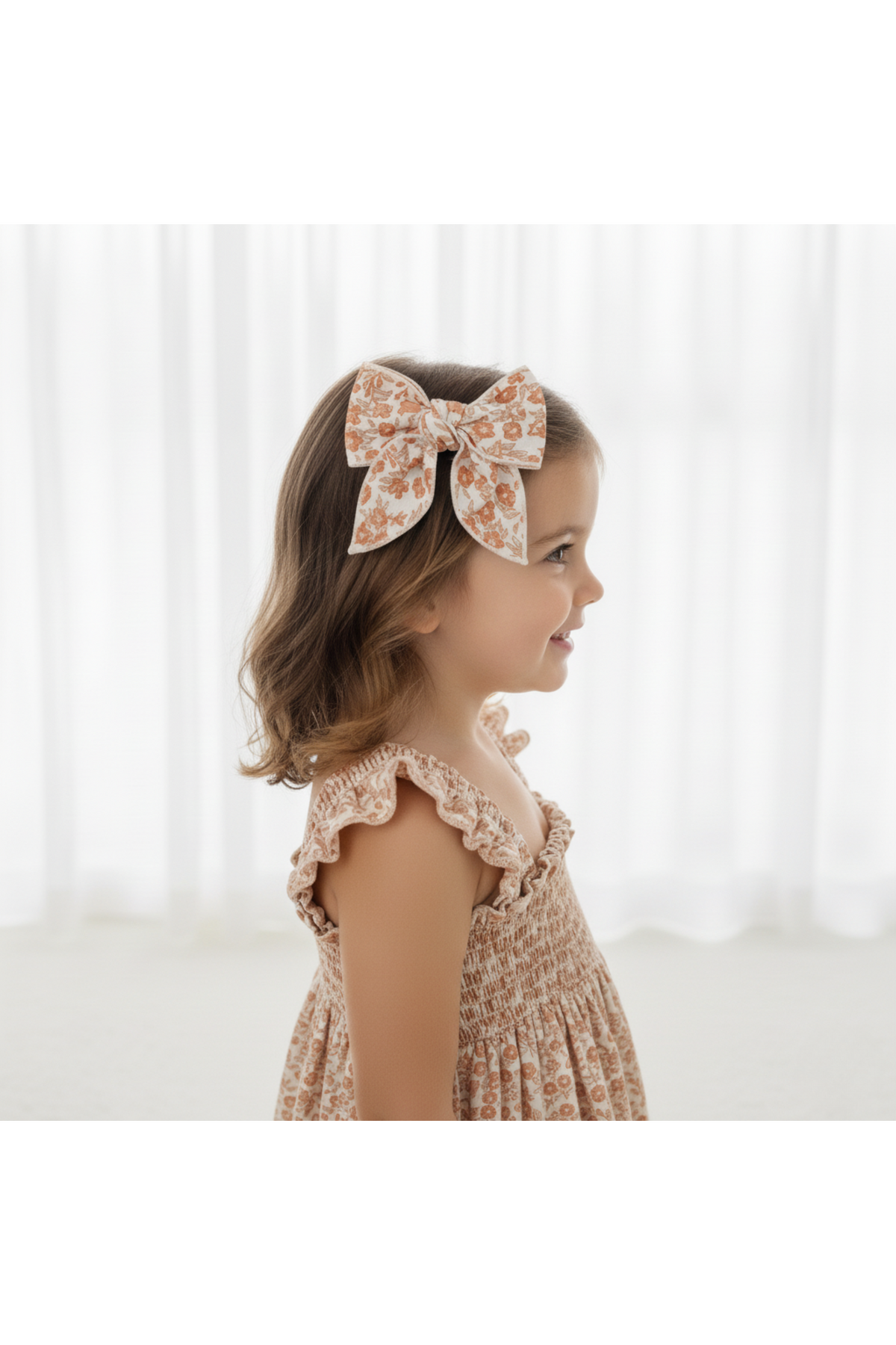A young girl with brown hair wears a floral-patterned hair bow clip in earthy tones. She is dressed in a smocked, light-colored dress and is posed against a bright, airy.