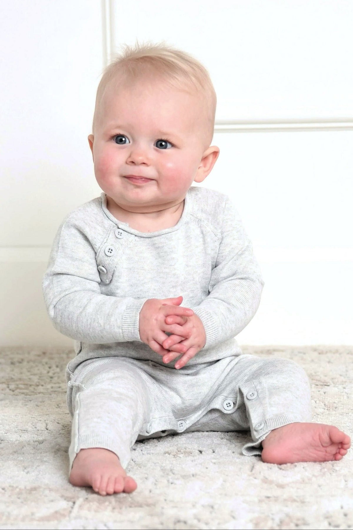 A baby sits on a soft rug, wearing a light gray organic cotton romper with button details and a cozy.