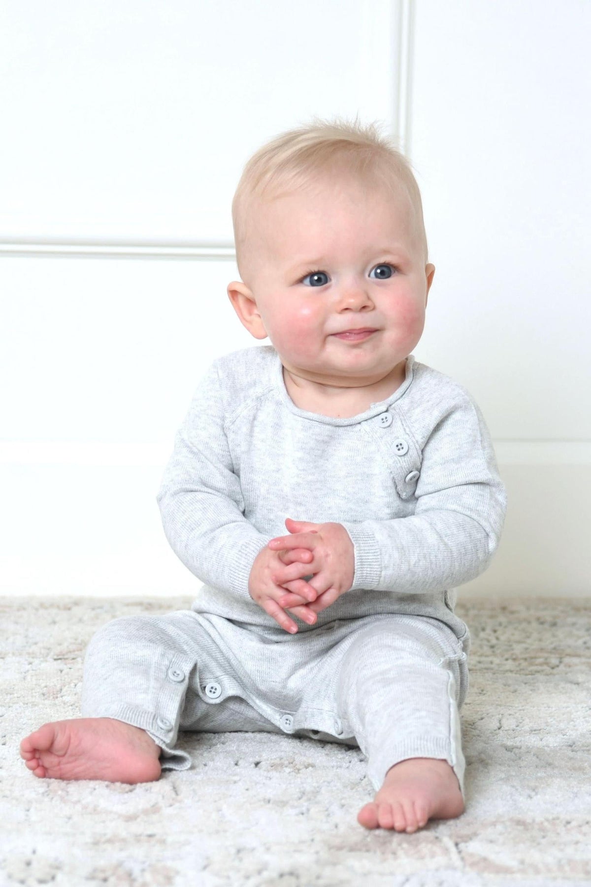 A smiling baby sits on a soft rug, wearing a light heather grey organic cotton romper with buttons at the shoulders and legs, showcasing a cozy and comfortable style.