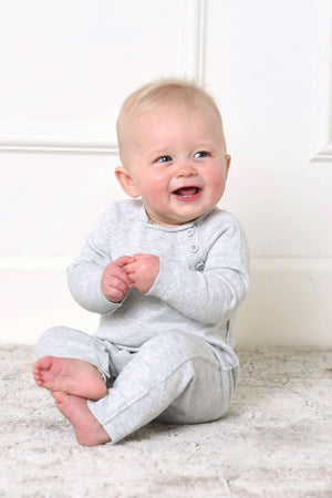 A smiling baby sits on a soft rug, wearing a light heather grey organic cotton romper with rolled edges and shoulder buttons. The cozy outfit highlights sustainable fashion for infants.