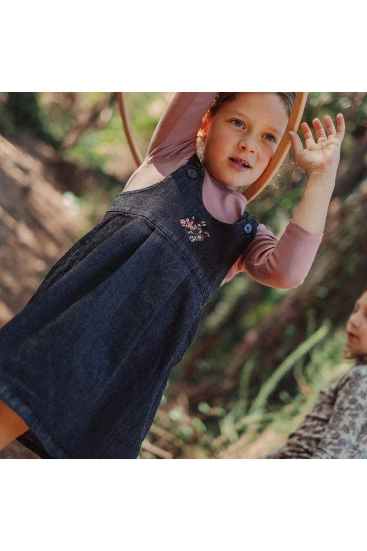 A young girl plays outdoors in a black denim dress, featuring delicate floral embroidery and adjustable straps. She is surrounded by greenery, showcasing a fun and active moment.