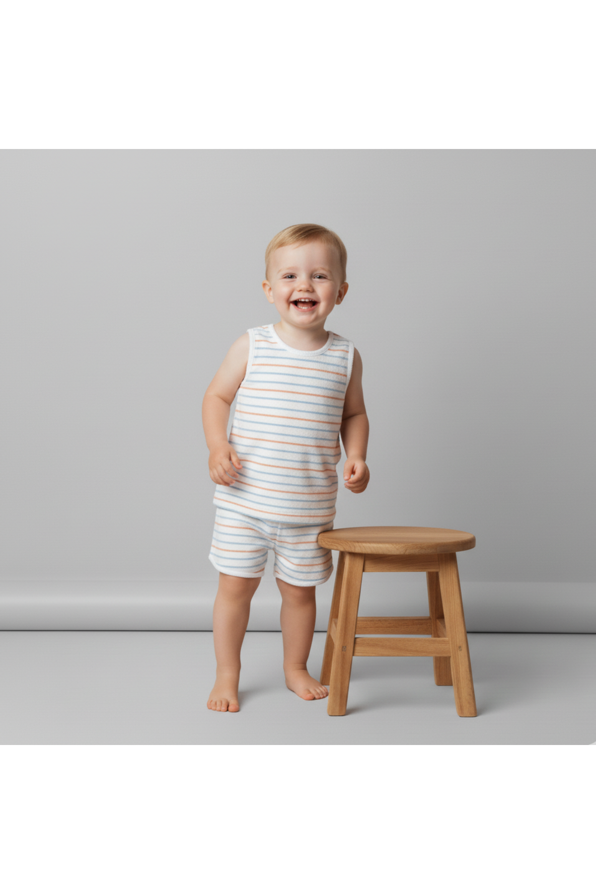 A smiling toddler wearing a striped organic cotton sleeveless tee and shorts stands beside a wooden stool against a neutral background, highlighting the relaxed and playful nature of the outfit.