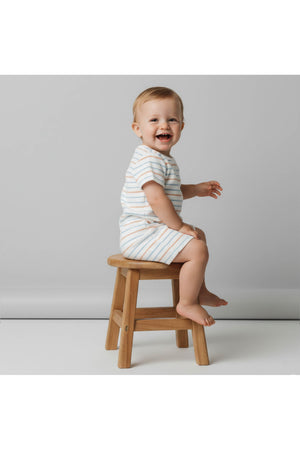 A smiling baby sits on a wooden stool, wearing a striped organic cotton romper in soft pastel colors. The neutral backdrop highlights the rompers coastal style and comfort.