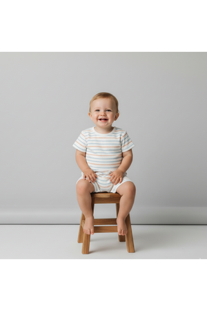A smiling toddler sits on a wooden stool, wearing a light-colored organic cotton short romper with subtle cabana stripes. The backdrop is neutral, enhancing the playful and relaxed vibe.