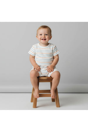 A smiling toddler sits on a wooden stool, wearing a short romper featuring light blue and white stripes. The outfit is made from soft organic cotton, perfect for warm weather.