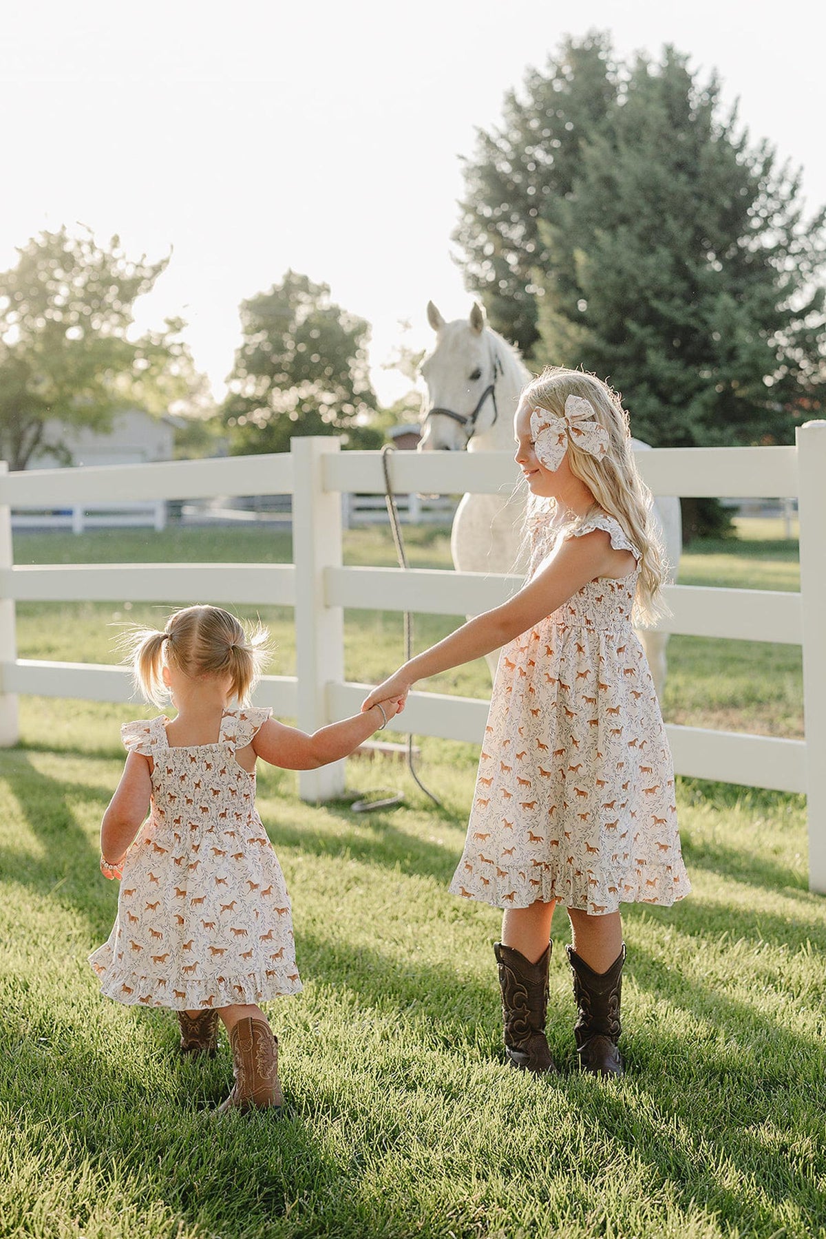 Two young girls in playful pony-printed dresses hold hands while standing in a grassy field, with a white horse nearby and a wooden fence in the background.