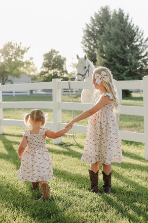 Two young girls in playful pony-printed dresses hold hands while standing in a grassy field, with a white horse nearby and a wooden fence in the background.