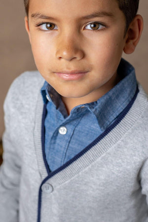A young boy is wearing a grey cardigan with navy tipping over a blue shirt, showcasing a stylish and refined.