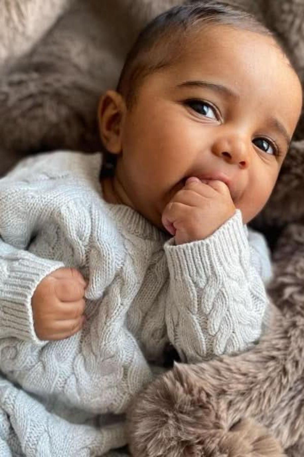 A baby wearing a light gray cable knit sweater romper looks curiously at the camera while sucking on their thumb, surrounded by a soft, plush blanket.