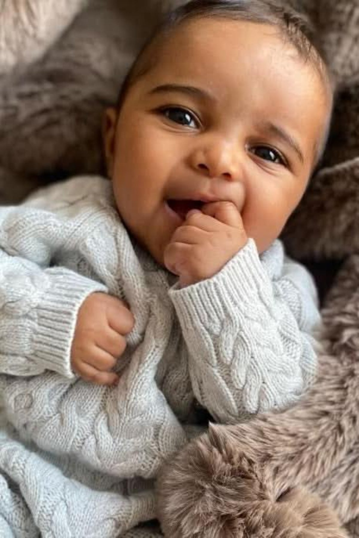 A smiling baby wearing a light gray cable knit sweater, made from soft organic cotton, sits on a plush, fur-like blanket. The baby is playfully sucking on their finger.