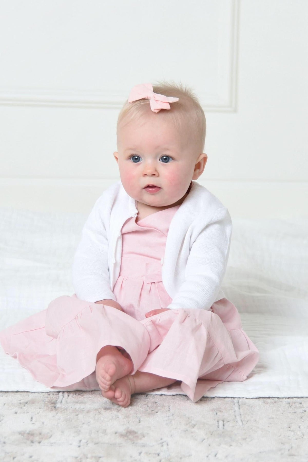 A baby girl sits on a soft surface, wearing a white cardigan over a pink dress, with a small bow.
