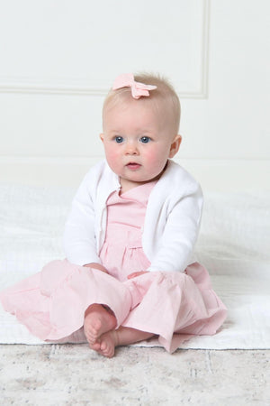 A baby girl sits on a soft surface, wearing a white cardigan over a pink dress, with a small bow.