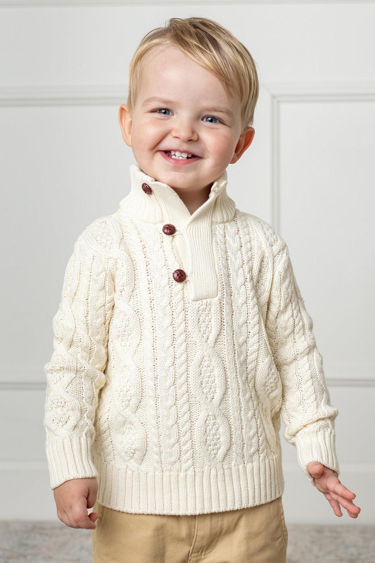 A smiling toddler wears a white organic cotton cable knit mock neck sweater featuring wooden buttons. The cozy sweater is perfect for keeping kids warm and stylish during cooler days.