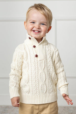 A smiling toddler wears a white organic cotton cable knit mock neck sweater featuring wooden buttons. The cozy sweater is perfect for keeping kids warm and stylish during cooler days.